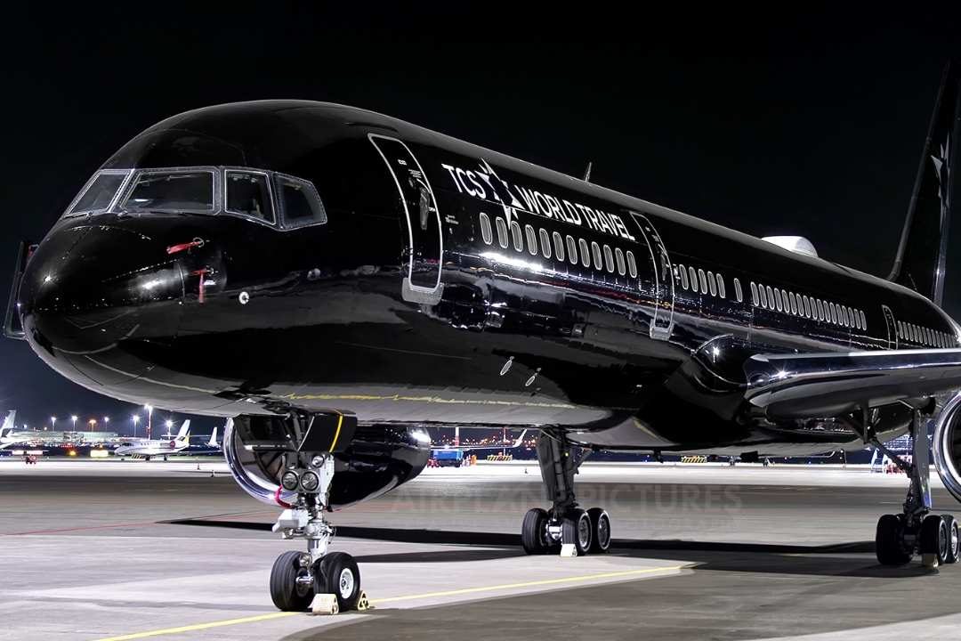 TCS World Travel A sleek black airplane with 'TCS World Travel' branding parked at night on an airport tarmac, illuminated by bright lights, showcasing its luxurious and modern design.